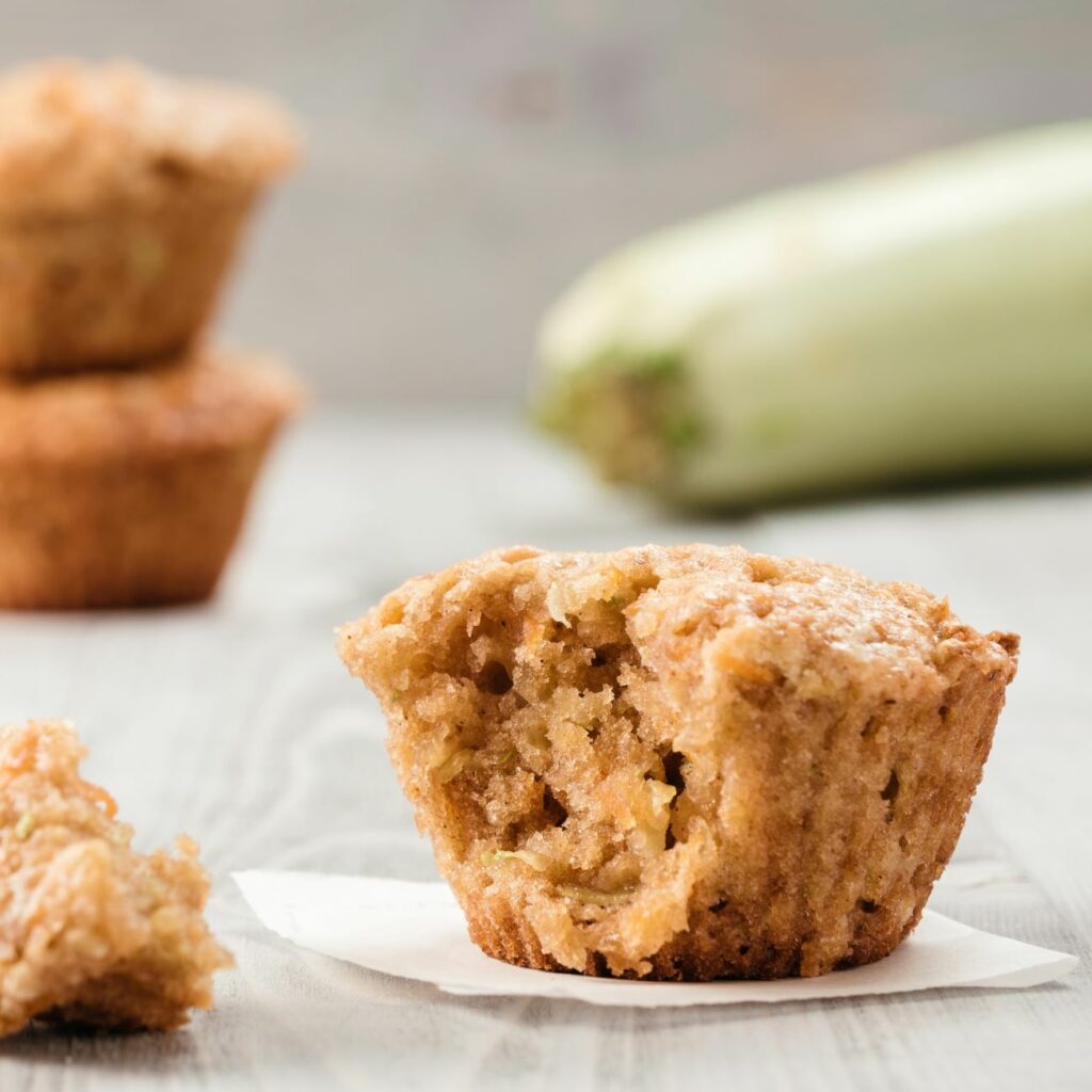 Zucchini-Karotten-Muffins werden angebissen mit einer Zucchini im Hintergrund präsentiert