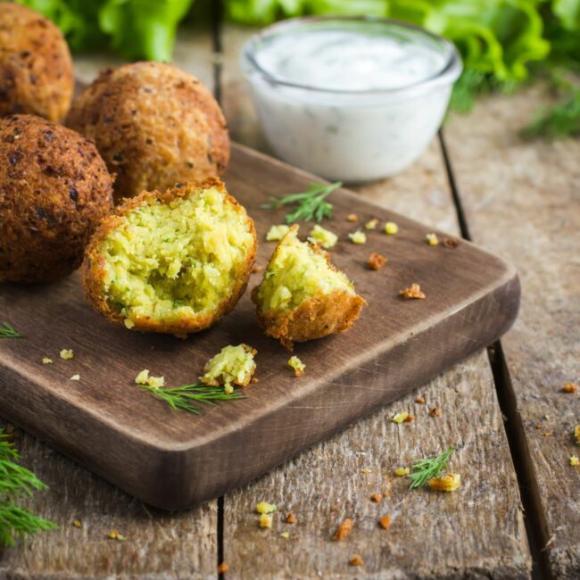 Falafel werden auf einem Holzbrett präsentiert, einer ist dabei angerissen.