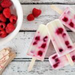 Himbeer-Eis am Stiel wird neben einer Schale frischer Himbeeren gezeigt.