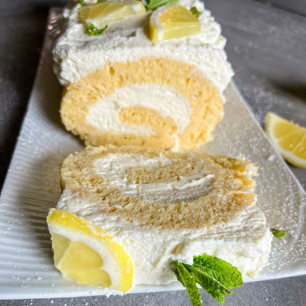 Zitronenrolle wird angerichtet auf einer weißen Tortenplatte gezeigt.