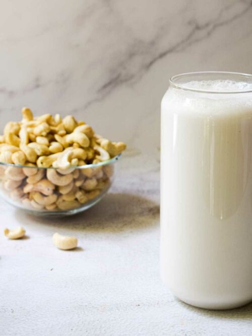 Cashewmilch wird in einem Glas vor einem Marmor-Hintergrund mit einer Schale Cashews gezeigt.