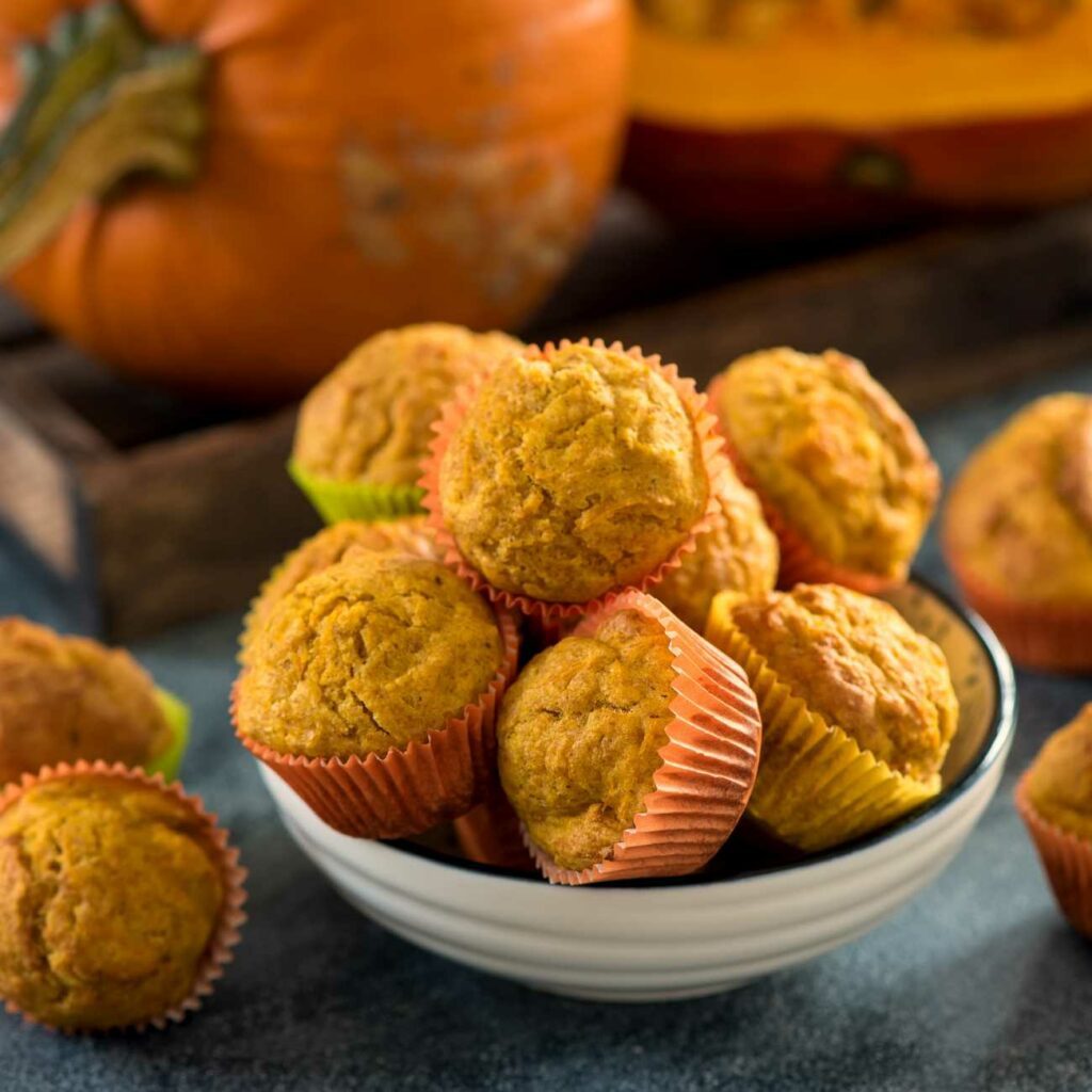 Tekvicové muffiny sú zobrazené naskladané v miske s tekvicami v pozadí.
