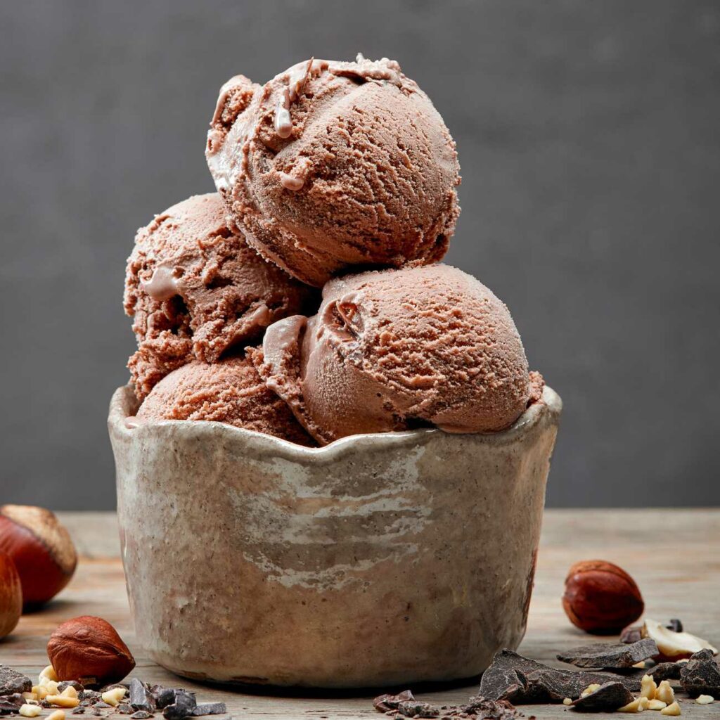 Lešnikov sladoled je prikazan v lončeni skledi z lešniki in nekaj čokolade.
