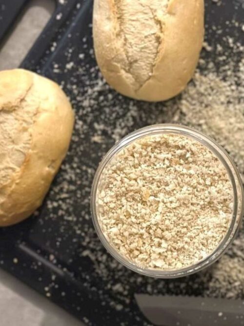 Semmelbrösel selber machen: Semmelbrösel werden in einem Glas auf einem Schneidebrett mit Brötchen gezeigt.